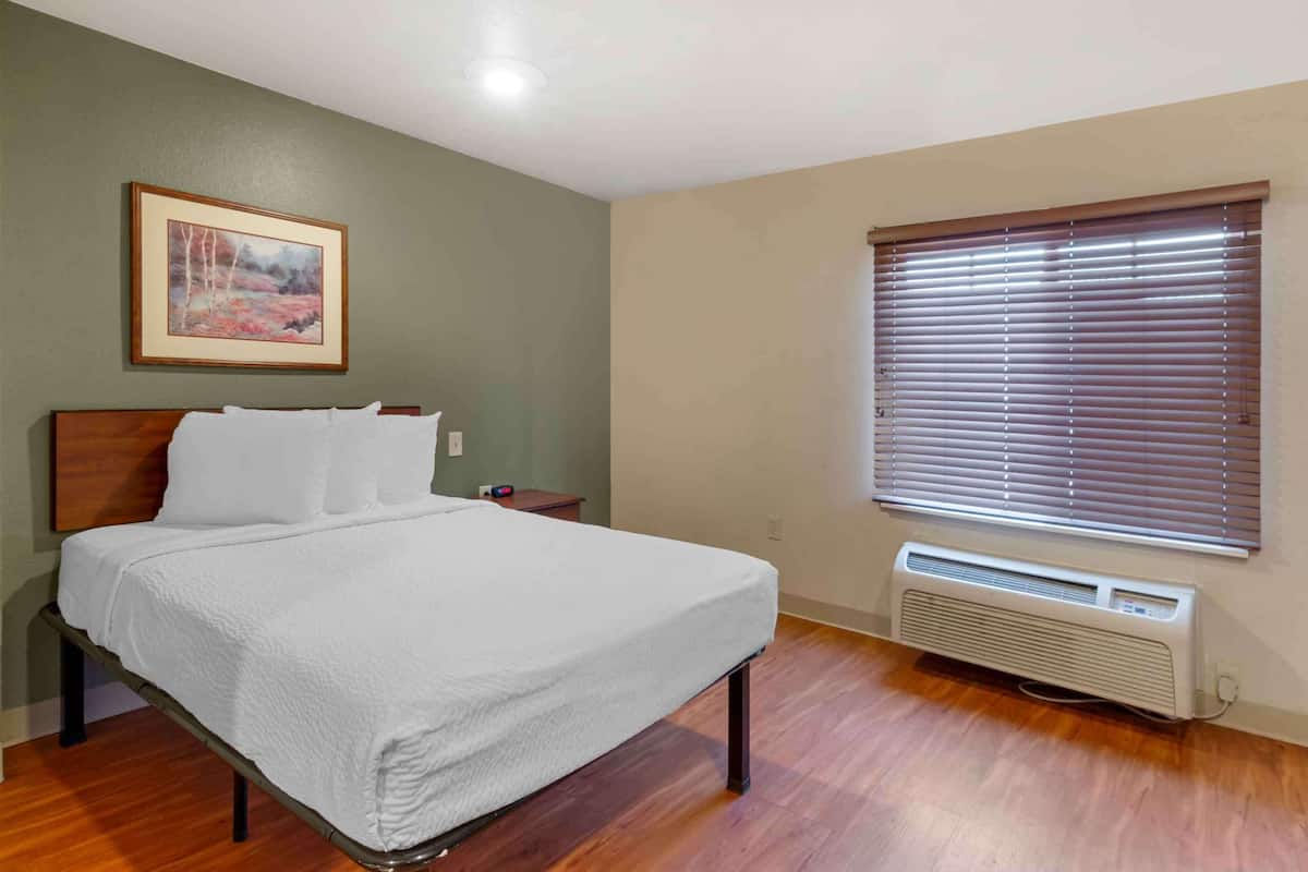 A neatly made bed with white linens in a room with green walls, a framed picture, a window with blinds, and a wall-mounted heater.