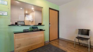 Uma mesa de recepção com um computador, uma placa indicando horários de check-in e check-out, uma placa que indica aceitação de animais de estimação e uma cadeira no canto.