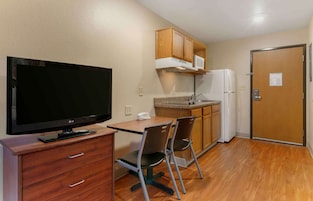 A compact kitchen with wooden cabinets, a microwave, a refrigerator, a dining table with two chairs, and a flat-screen TV on a wooden cabinet.
