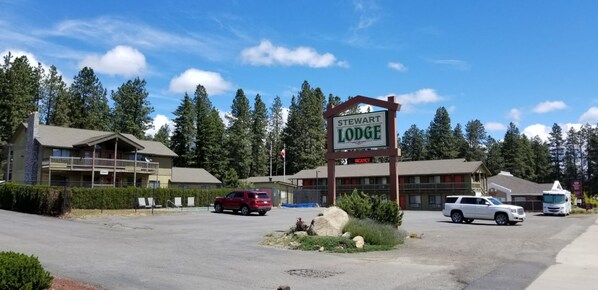 Señal de Stewart Lodge con un cartel de 'VACANCY' debajo, rodeada por un estacionamiento con una SUV roja y una SUV blanca, y una casa rodante blanca al fondo.