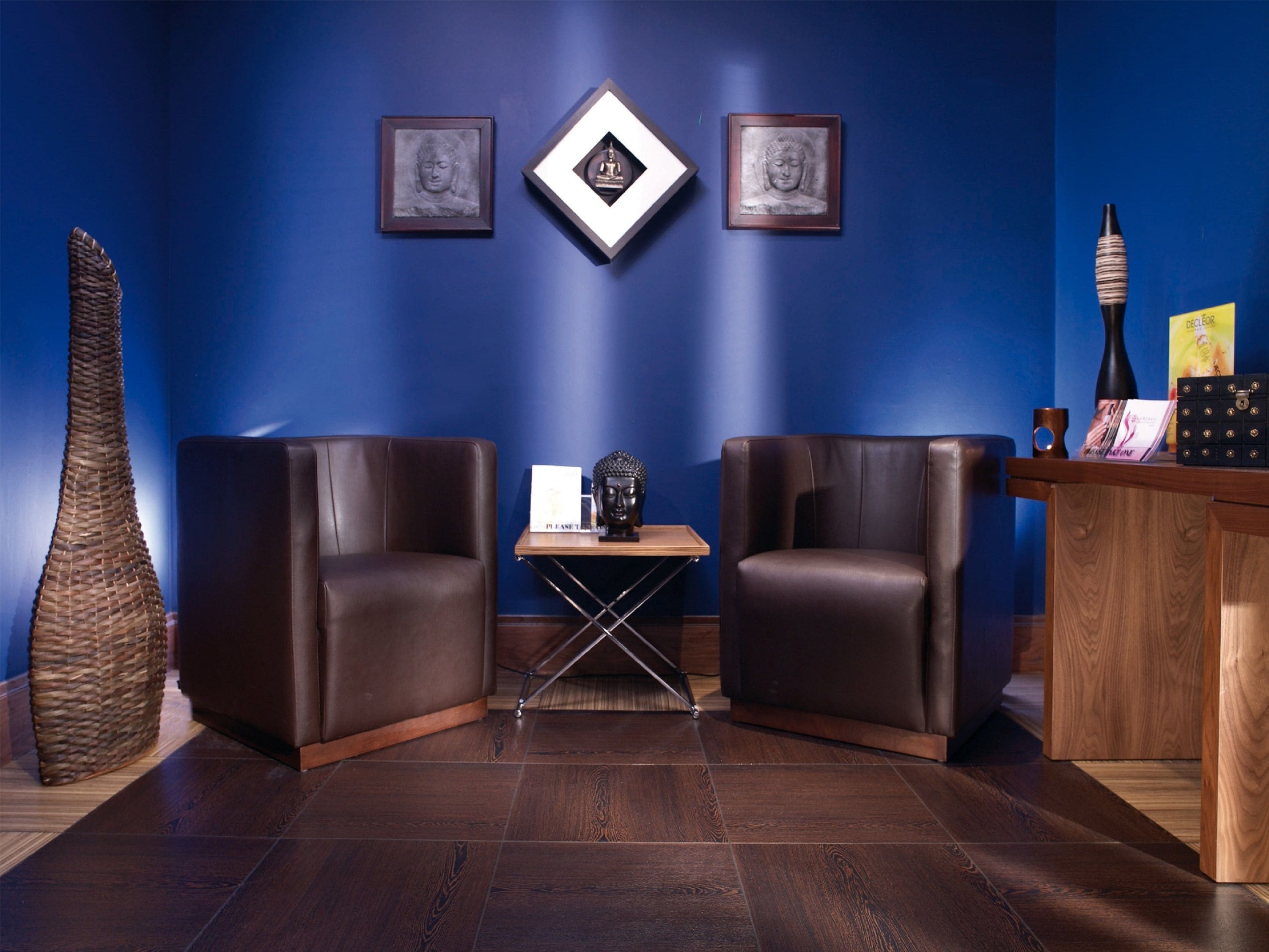 A room with two leather chairs, a small table with a Buddha statue, framed artwork on the wall, and a wooden desk with a vase and a book.