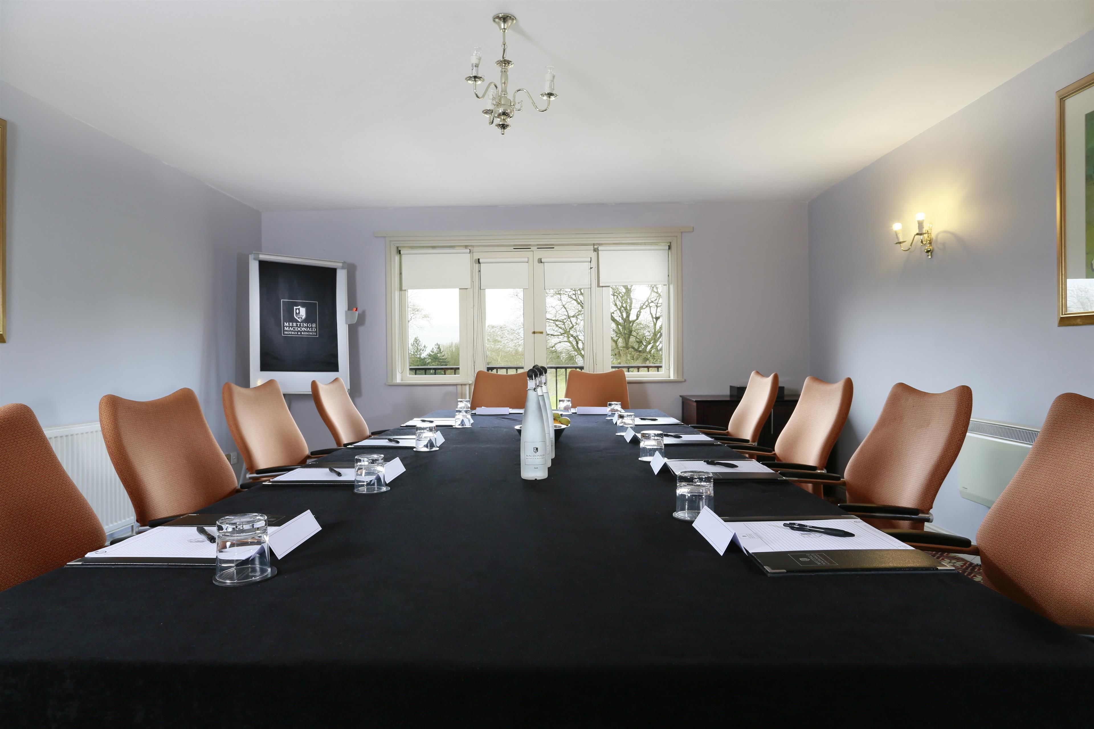 Una sala conferenze con un lungo tavolo, sedie marroni, un lampadario e una finestra con vista sugli alberi.