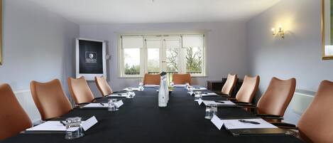 A conference room with a long table, brown chairs, a chandelier, and a window with a view of trees.