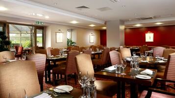 A well-lit dining area with multiple tables and chairs, a fire exit sign, and framed artwork on the walls.