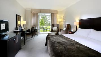 A hotel room with a bed, a desk with a television, a chair, a table with chairs, and a balcony with a view of trees.