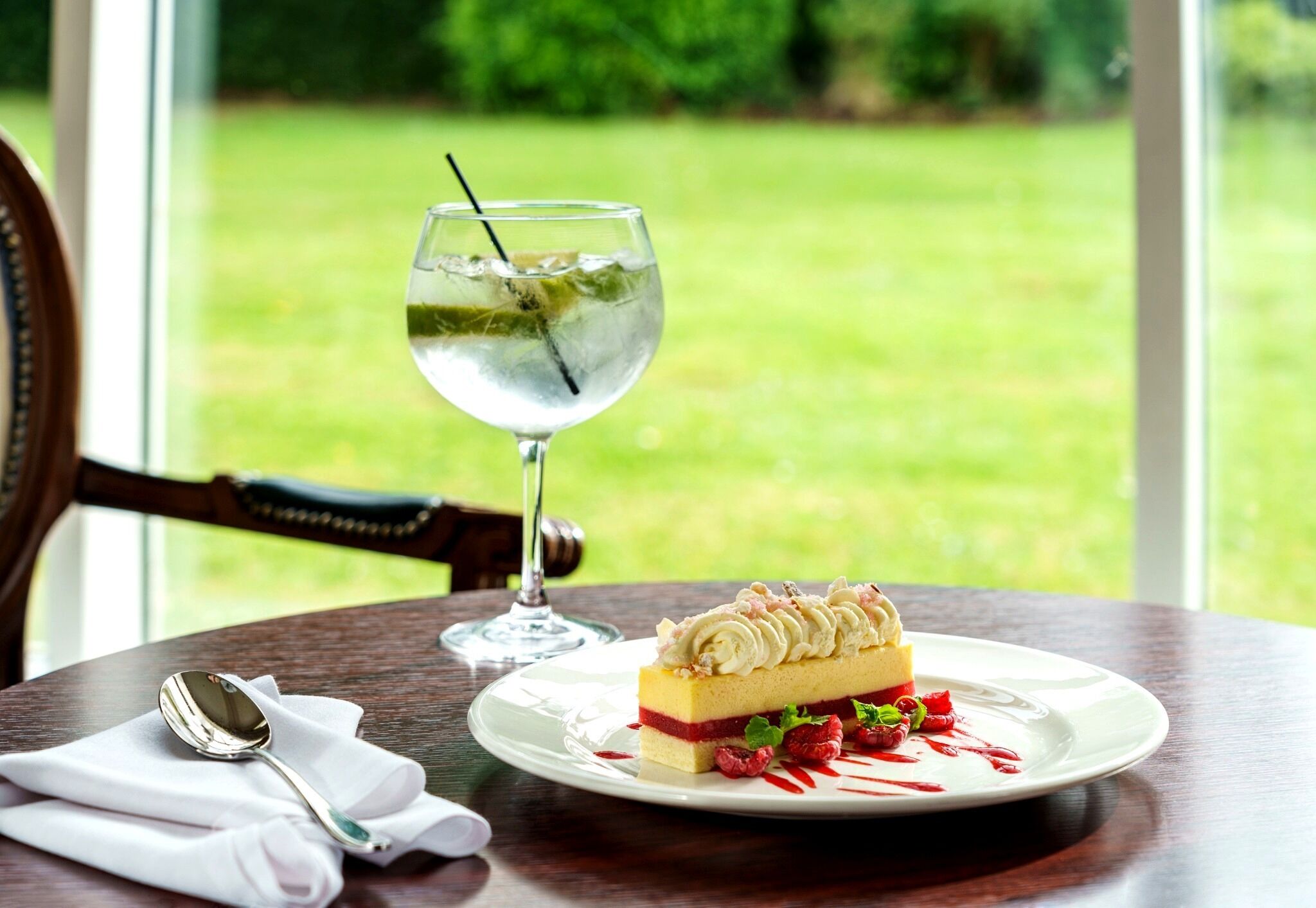 A dessert plate with a slice of cake, a glass of water, and a spoon on a table with a view of a grassy area.