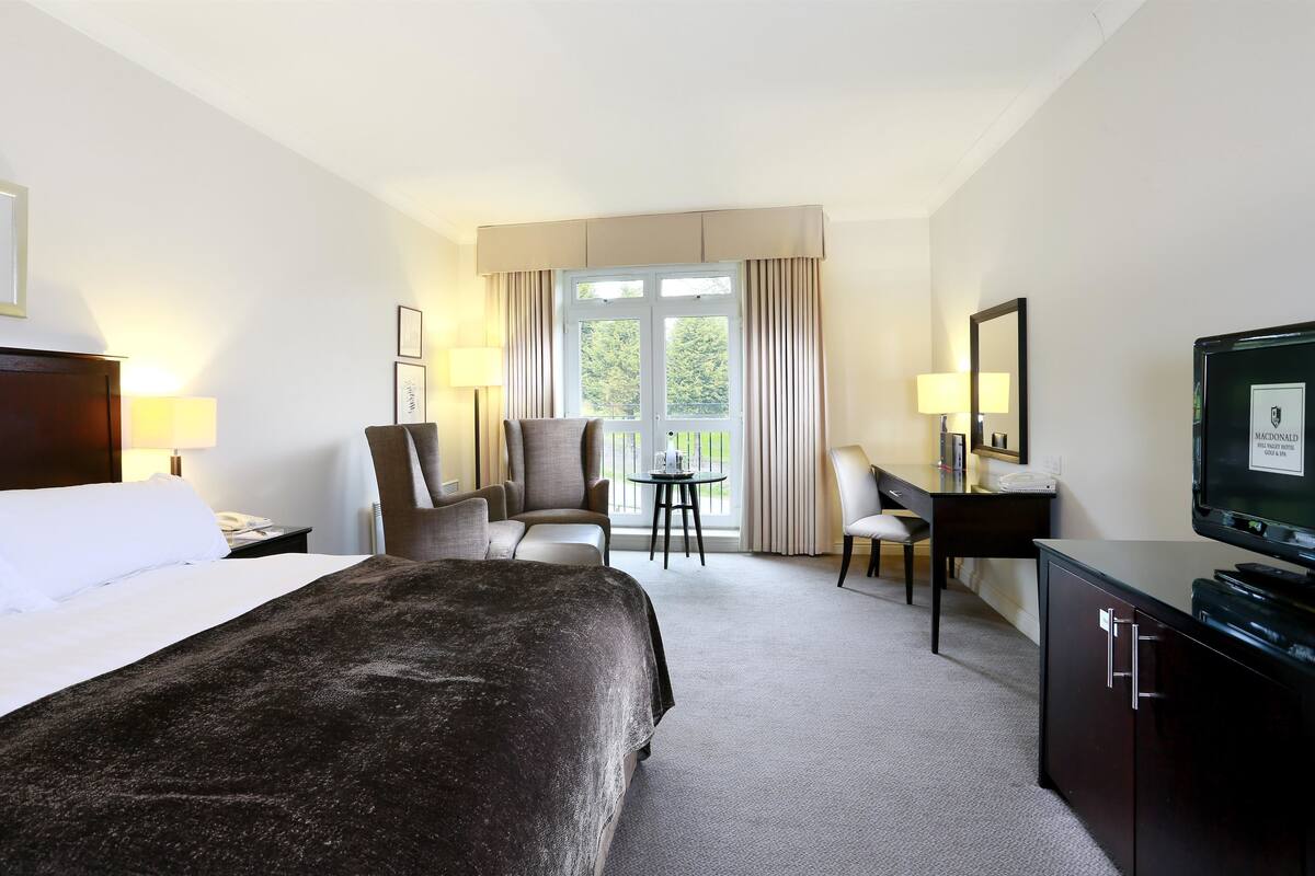 A hotel room with a large bed, a desk with a chair, a television, and a balcony with curtains.