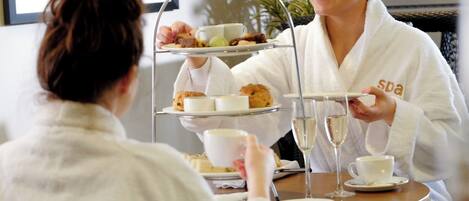 A person in a white robe serving a three-tiered tray of food and drinks to another person at a table.