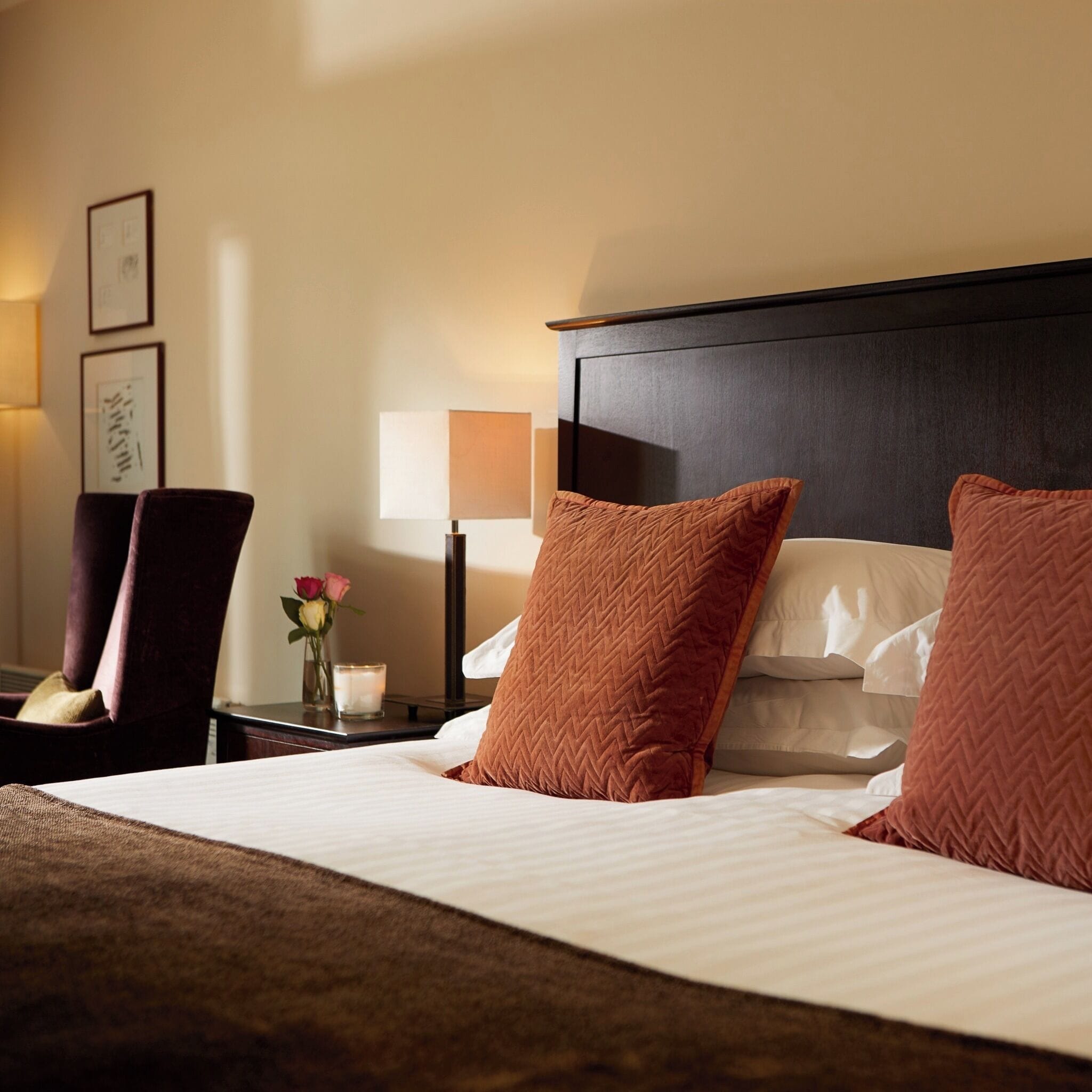 Habitación de hotel con cama, almohadas, mesita de noche, lámpara, sillón y cuadros en la pared.