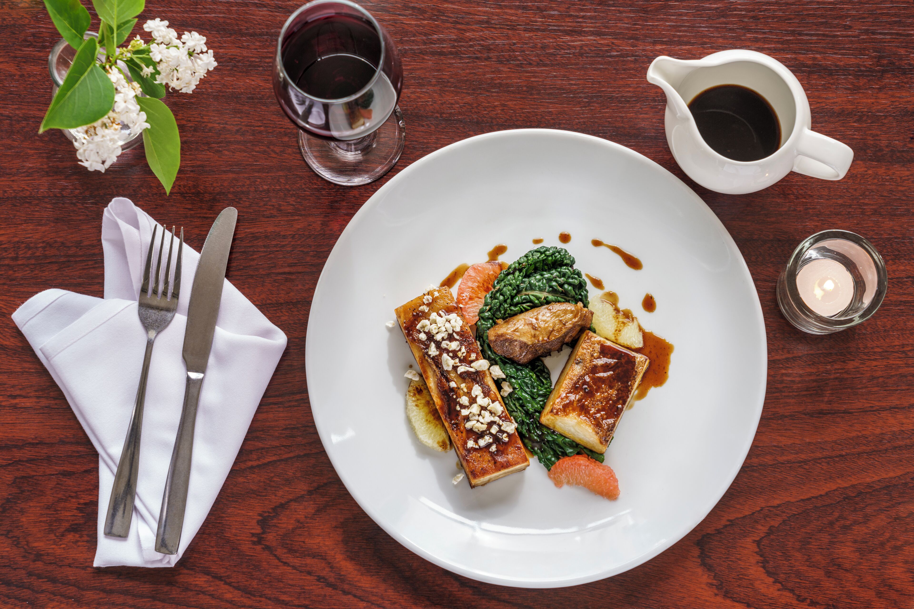 A wooden table with a white plate of food, a fork and knife, a small vase with flowers, and two glasses of a dark liquid.
