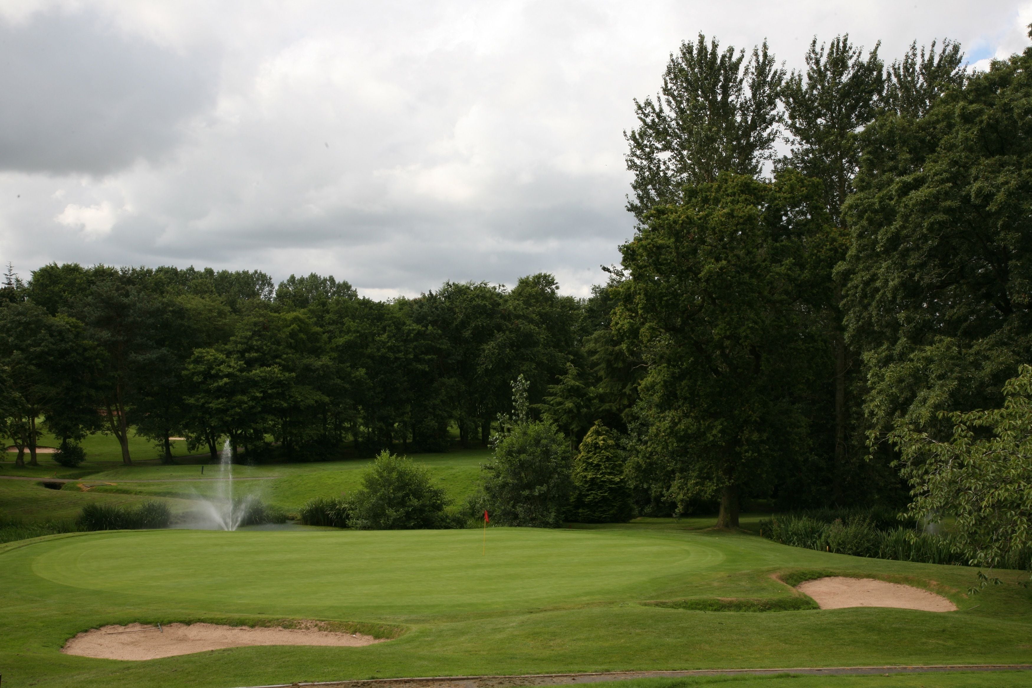 A golf course with a water fountain, sand bunkers, and dense trees in the background.