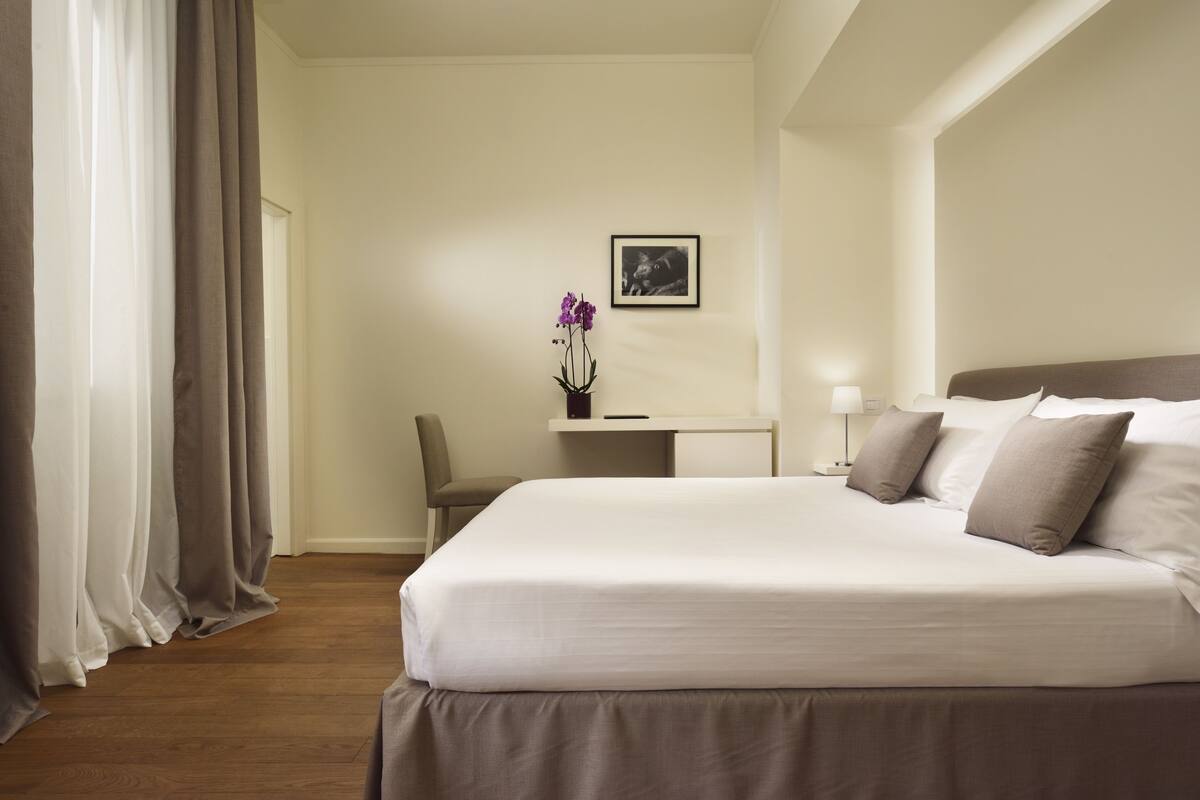 Une chambre d’hôtel avec un grand lit, un bureau avec une chaise, une petite table sur laquelle est posé un vase de fleurs, et un tableau encadré au mur.
