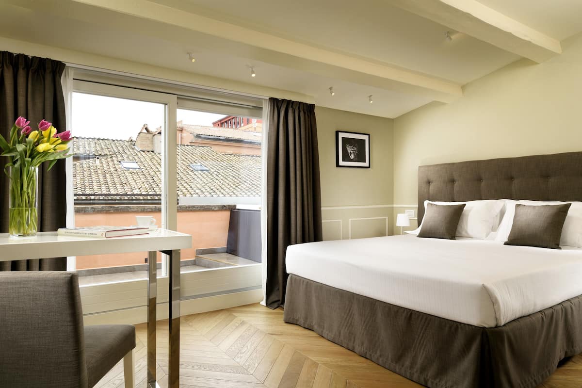 Une chambre d’hôtel avec un grand lit, une fenêtre avec des rideaux, une petite table sur laquelle est posé un vase de fleurs, et une vue sur le paysage urbain.