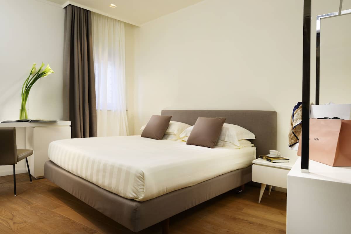 Une chambre d’hôtel moderne avec un grand lit, un bureau, une chaise, une table de chevet avec un livre et une tasse, et un grand miroir.