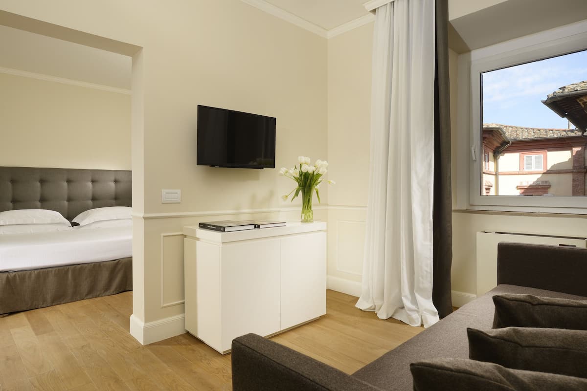 Une chambre d’hôtel avec un lit, un téléviseur fixé au mur, un petit bureau blanc orné d’un vase de fleurs et une vue sur l’extérieur par une fenêtre.