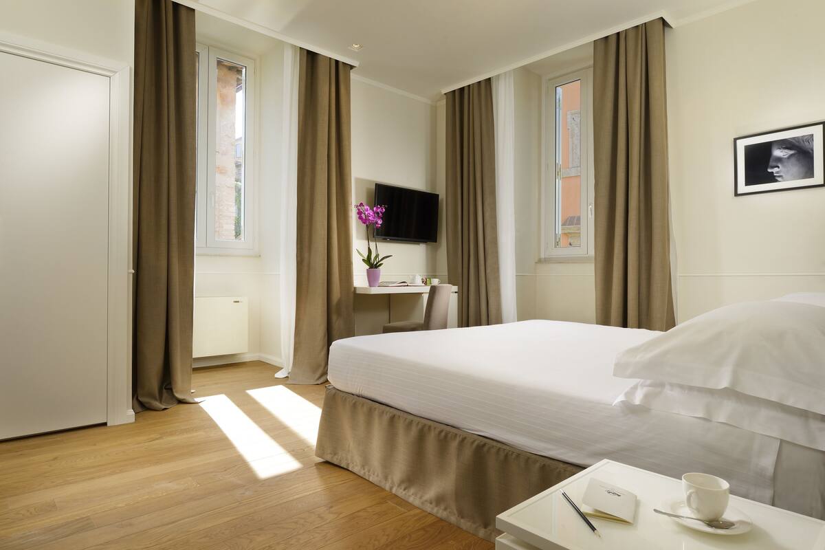 Une chambre d’hôtel avec un lit, un bureau avec une tasse et un livre, un téléviseur fixé au mur et des rideaux.