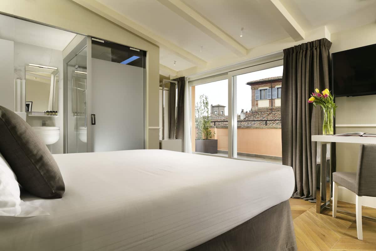 Une chambre d’hôtel moderne dotée d’un grand lit, d’un bureau avec une chaise, d’une télévision à écran plat et d’un balcon offrant une vue sur les bâtiments.