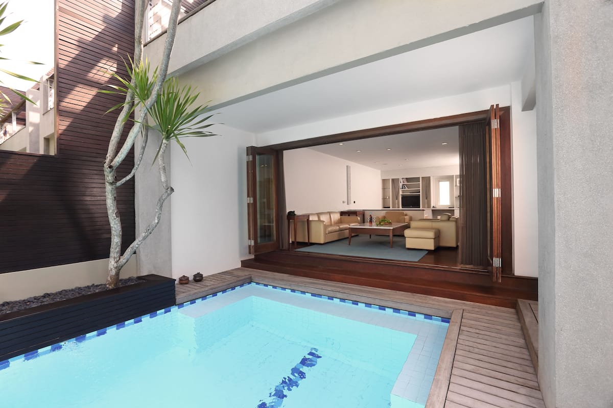 A modern outdoor pool area with a wooden deck, a built-in planter with a tree, and a view into a living room with a sofa and a coffee table.
