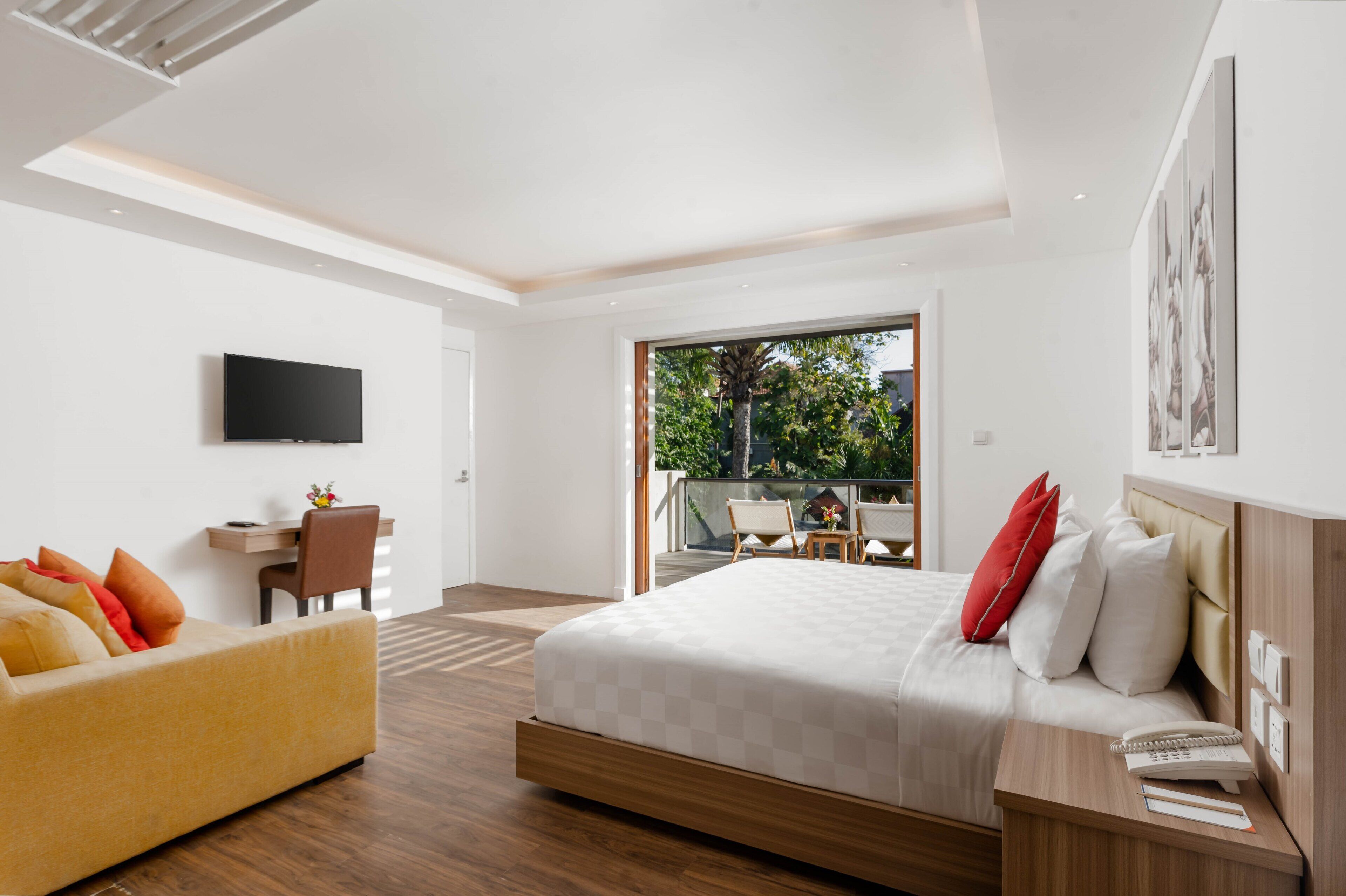 A modern hotel room with a bed, a desk, a sofa, and a balcony with outdoor seating.