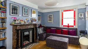 A living room with a fireplace, a sofa, a bookshelf, and a window with red curtains. - Brightonwave (Brighton)