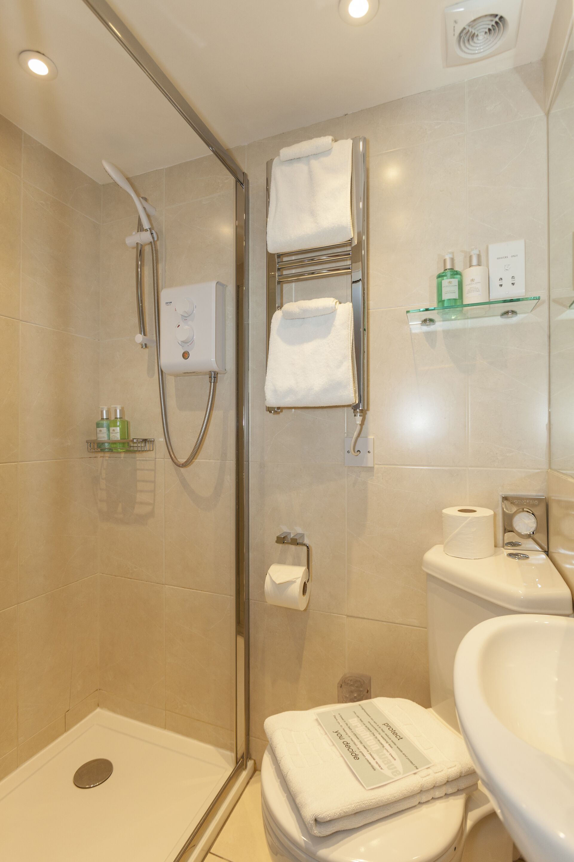 A bathroom with a glass-enclosed shower, a toilet, a sink, and a glass shelf with toiletries.