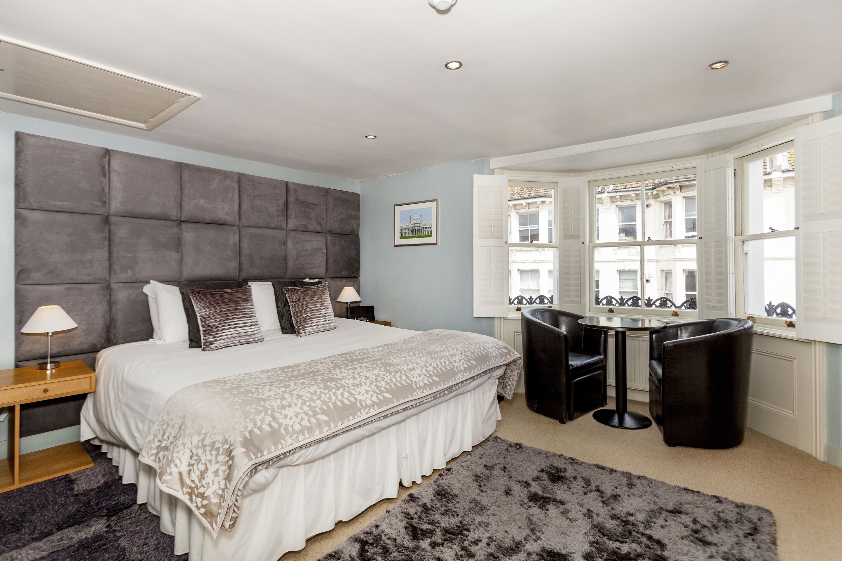 A bedroom with a large bed, a bedside table with a lamp, a small round table with two chairs, and a window with a view of buildings.