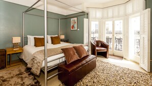A four-poster bed with a brown leather bench, a nightstand with a lamp, and a view of the city through large windows. - Brightonwave (Brighton)