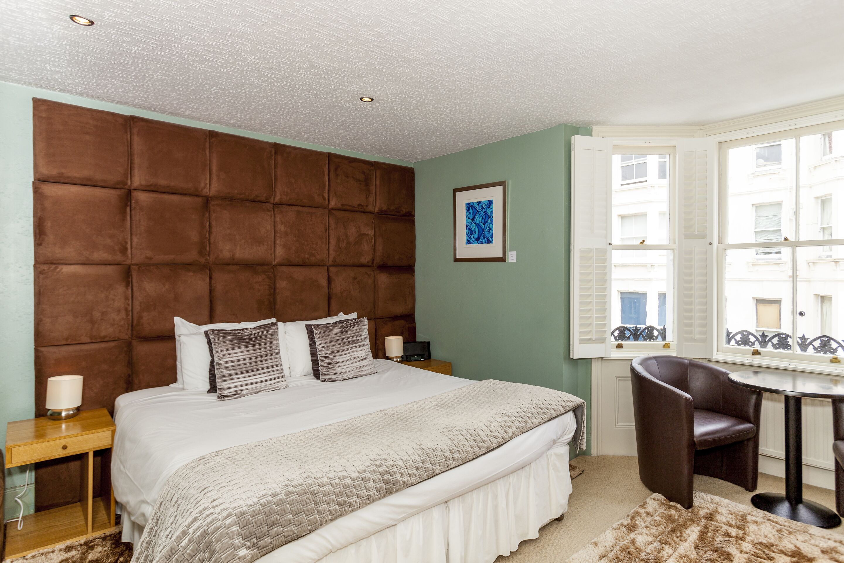 Ein Schlafzimmer mit einem großen Bett, einem Kopfteil, einem Nachttisch, einem Sessel, einem Fenster und einem gerahmten Bild an der Wand.