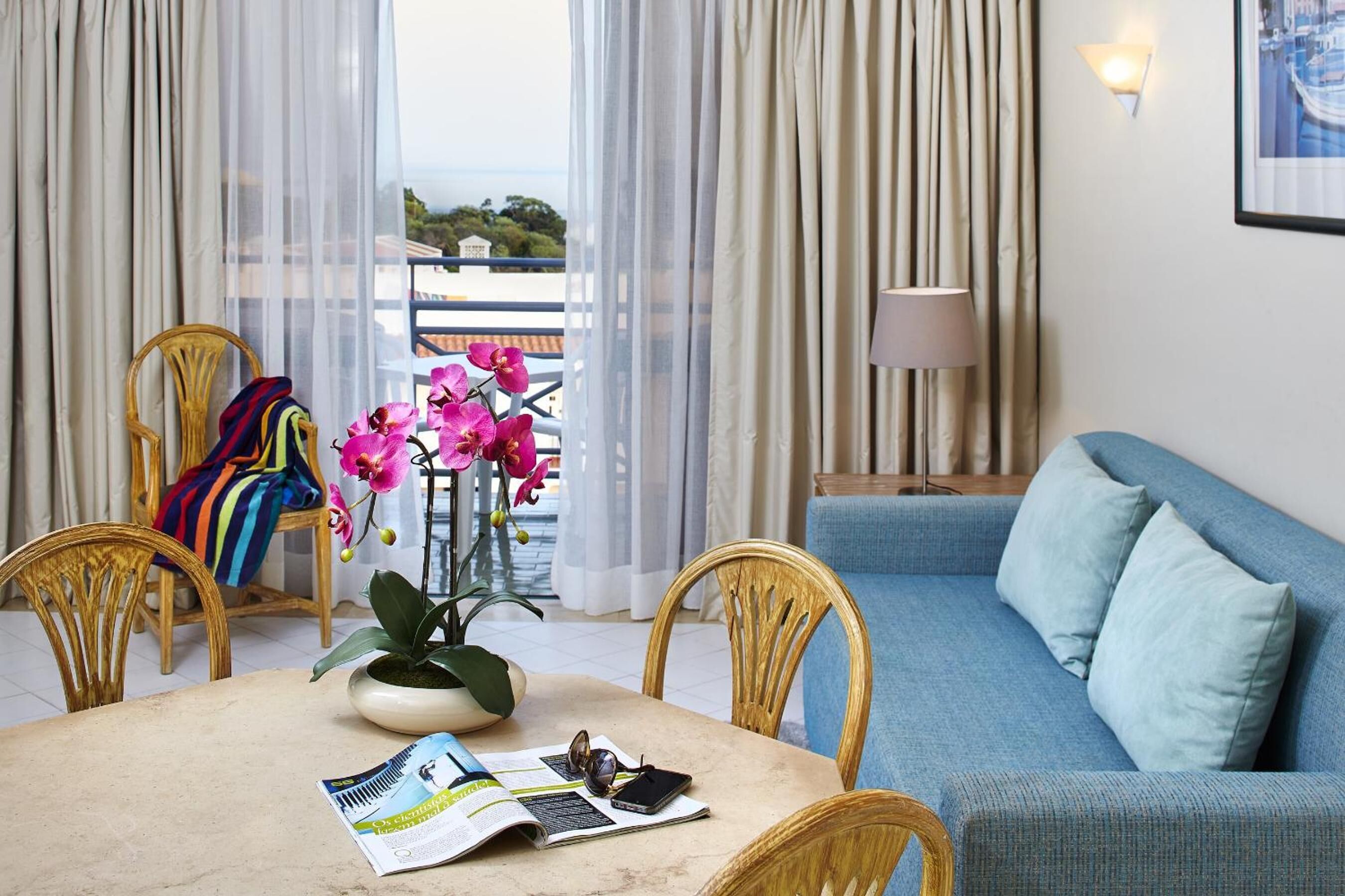 Une pièce avec un canapé bleu, une table à manger entourée de chaises, une orchidée en pot et une vue sur l’extérieur à travers des rideaux légers.