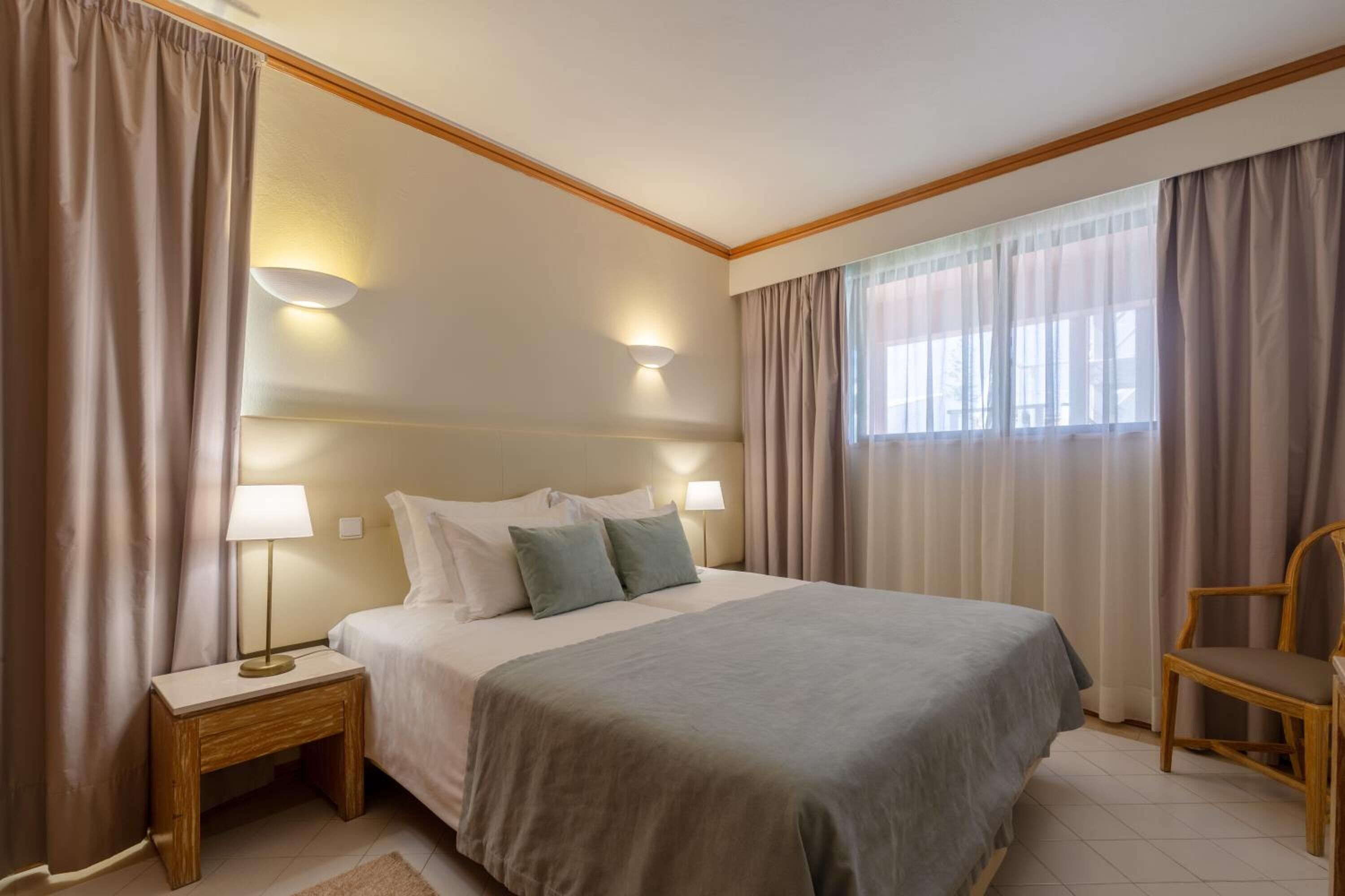 A neatly arranged hotel room with a large bed, two bedside tables with lamps, a chair, and a window with curtains.