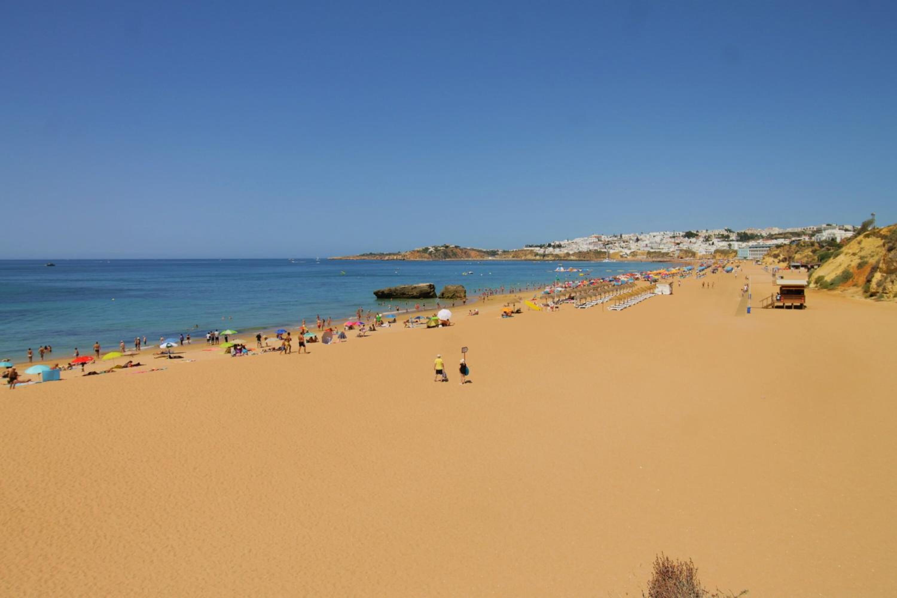 A wide sandy beach with people and umbrellas, a clear blue sky, and buildings in the background.