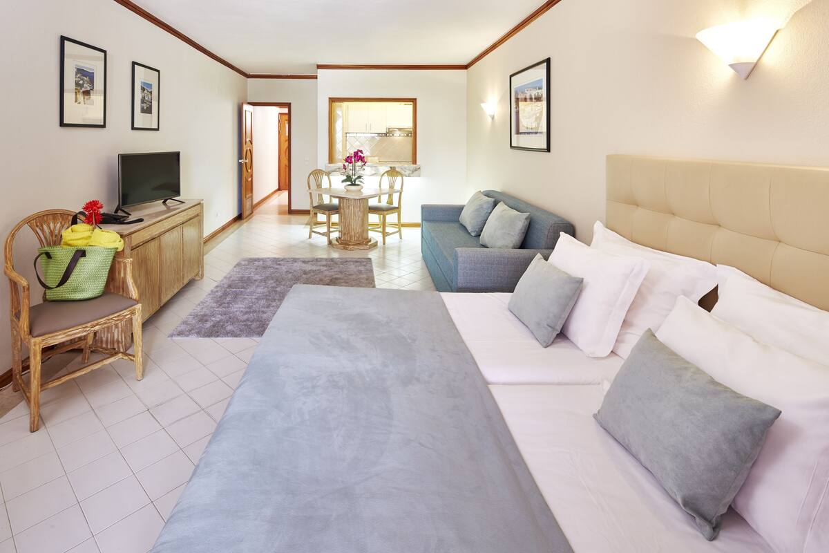 A hotel room with a bed, a sofa, a TV, a dining table with chairs, and a kitchen area in the background.