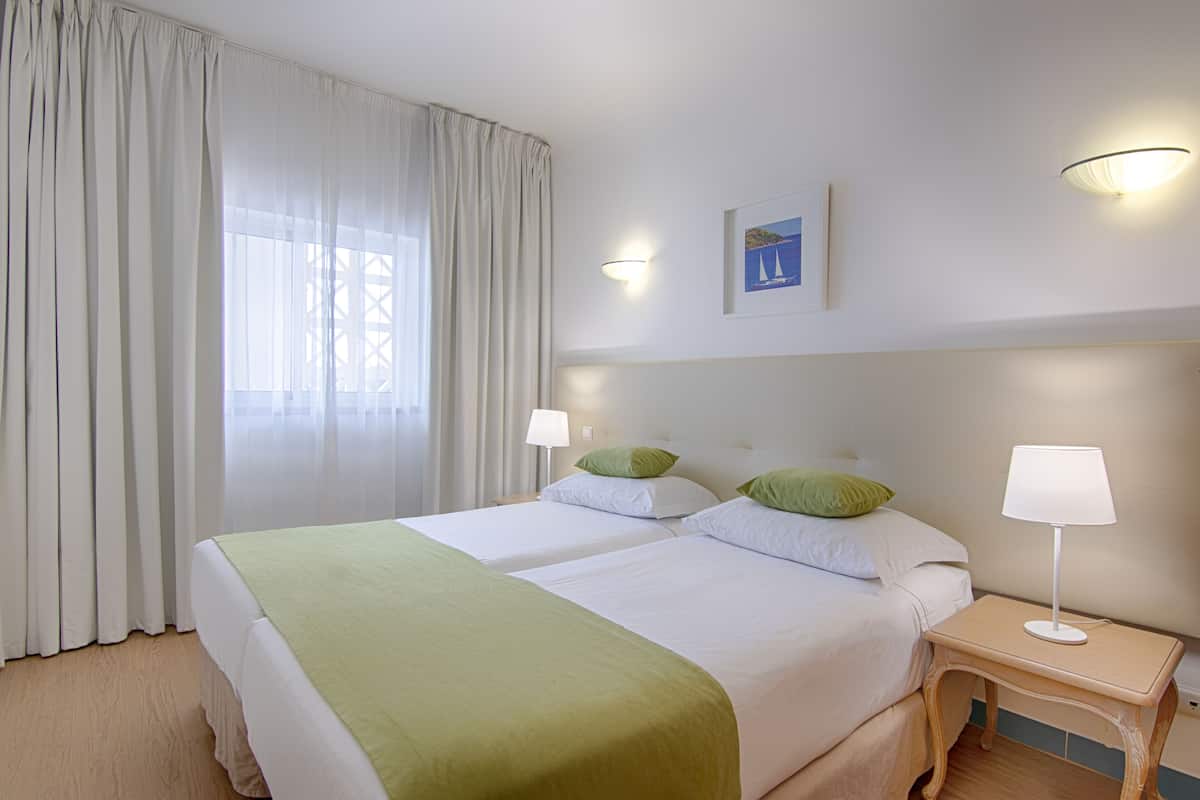 A hotel room with two beds, a nightstand, a lamp, a painting, and sheer curtains.