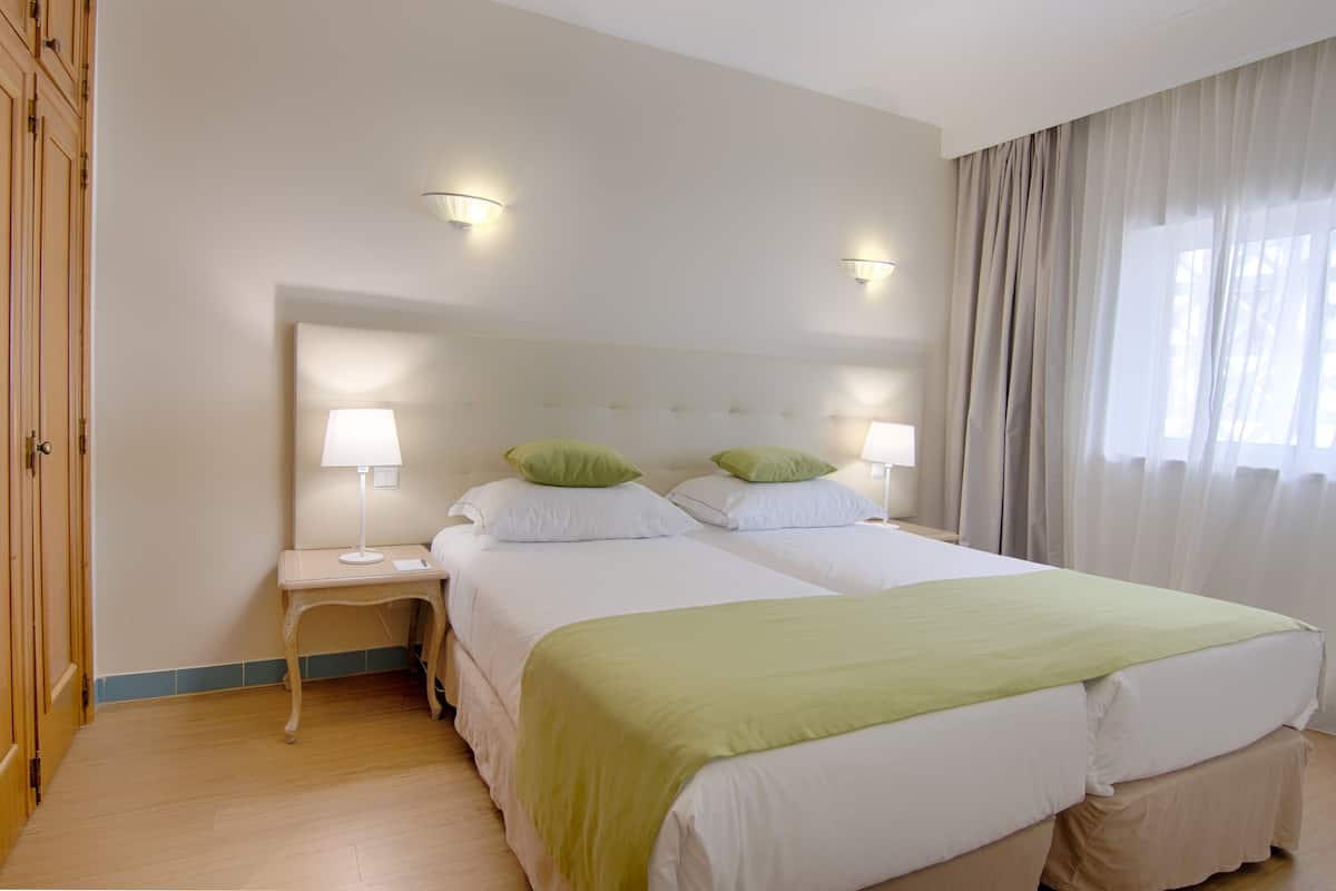 A hotel room with two beds, a nightstand, a lamp, and a window with curtains.
