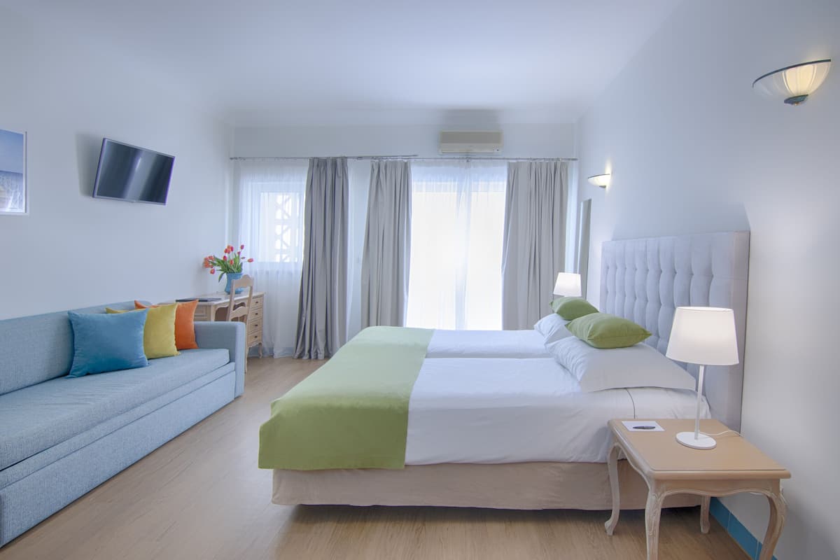 A hotel room with a bed, a sofa, a bedside table with a lamp, a TV on the wall, and curtains.