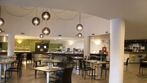 Een ruim restaurant met hoge tafels en stoelen, hangende吊り下げ式灯具, en een centraal gelegen toonbank met een groene accentmuur.