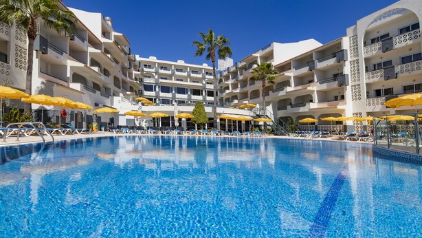 Een hotel met een centraal gelegen zwembad, meerdere balkons en gele parasols.