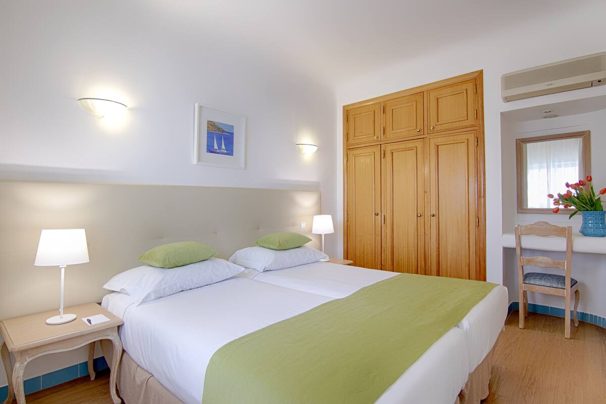 A double bed with green bedding, a nightstand with a lamp, a wooden wardrobe, and a chair with a vase of flowers.