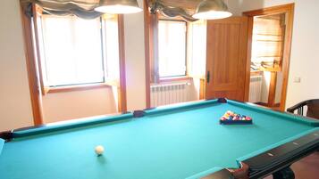 A pool table with a cue ball and a set of billiard balls, two pendant lights, and a window with curtains.