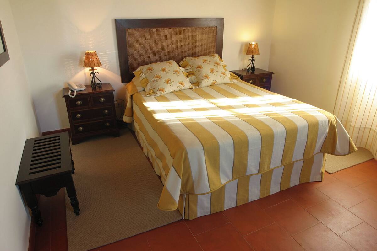 A bed with a striped bedspread, two bedside lamps, a nightstand with a phone, and a small table with a vent.