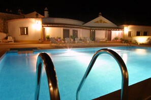 Una piscina con barandillas metálicas, rodeada de sillas de descanso y plantas en macetas, con un edificio de techo con tejas al fondo.