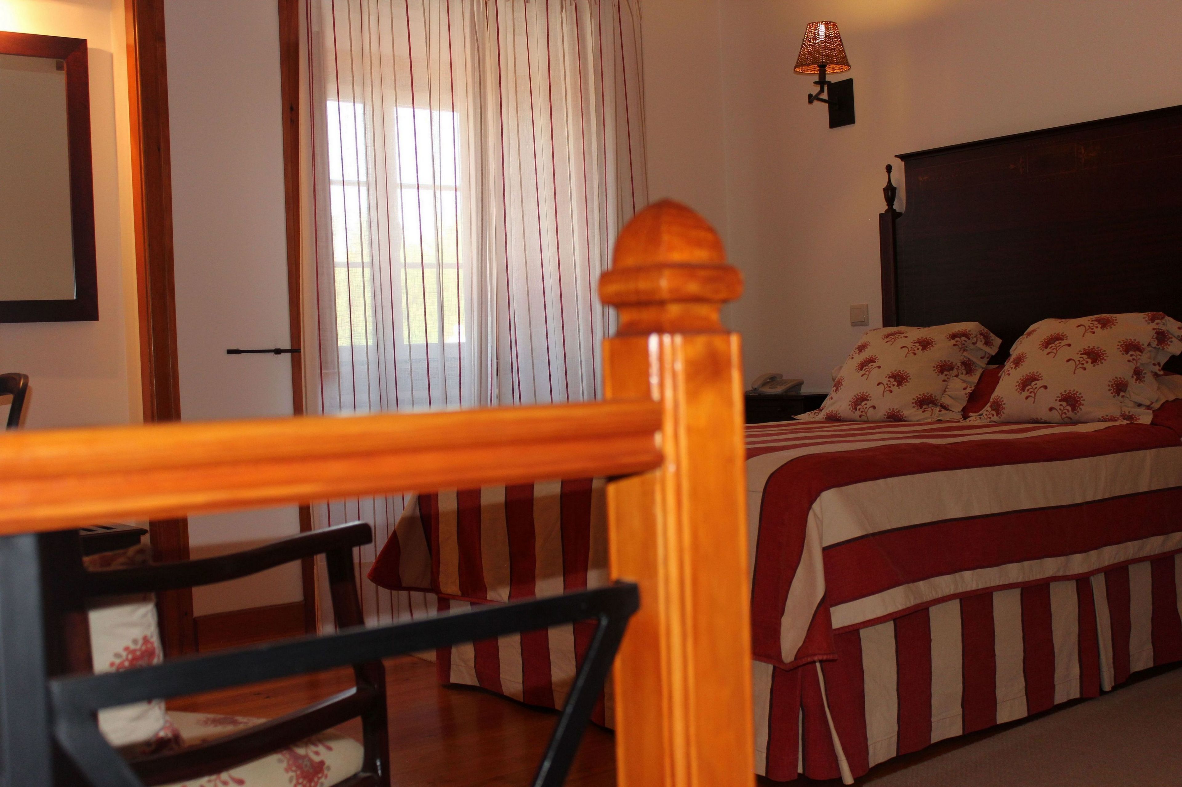 A hotel room with a bed, a chair, a wooden headboard, a window with curtains, and a wall-mounted lamp.