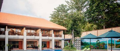 A swimming pool with a curved edge, surrounded by lounge chairs and umbrellas, and a building with a tiled roof and balconies.