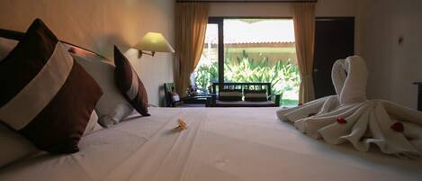 Habitación de hotel con cama, mesita de noche, lámpara y vistas a un balcón con plantas.