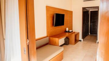 A hotel room with a wooden bench, a flat-screen TV mounted on the wall, a small table with a remote control, and a trash bin.