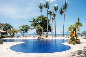 Una piscina con sillones de descanso y palmeras, rodeada de un área pavimentada y sombrillas.