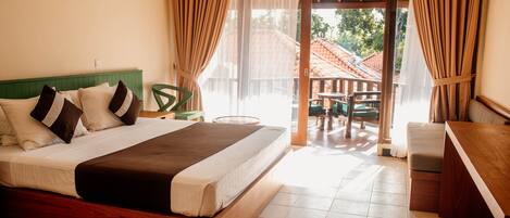 A hotel room with a large bed, a wooden desk, a balcony with a table and chairs, and a view of the outside.