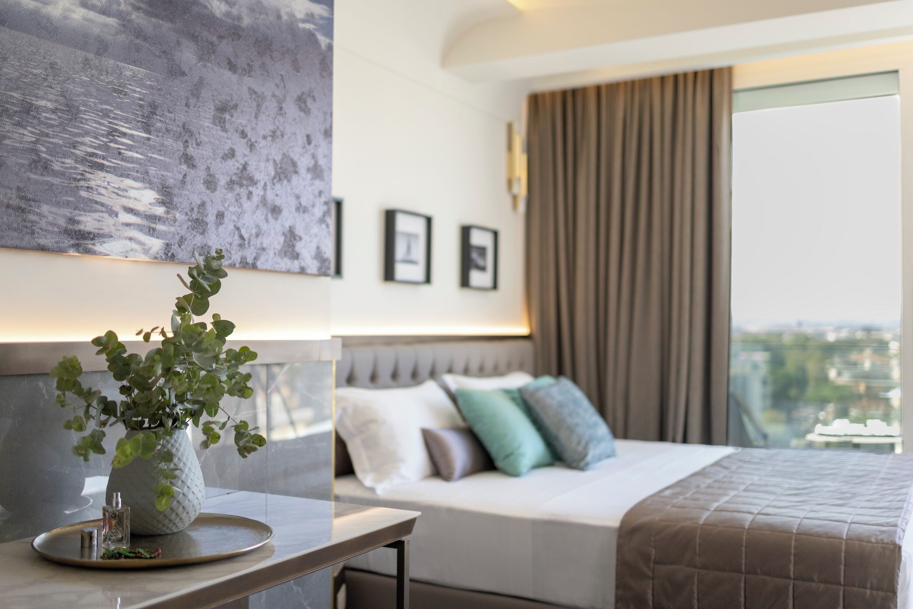 Una habitación de hotel moderna con una cama, un florero con una planta, cuadros en la pared y vistas a la ciudad.