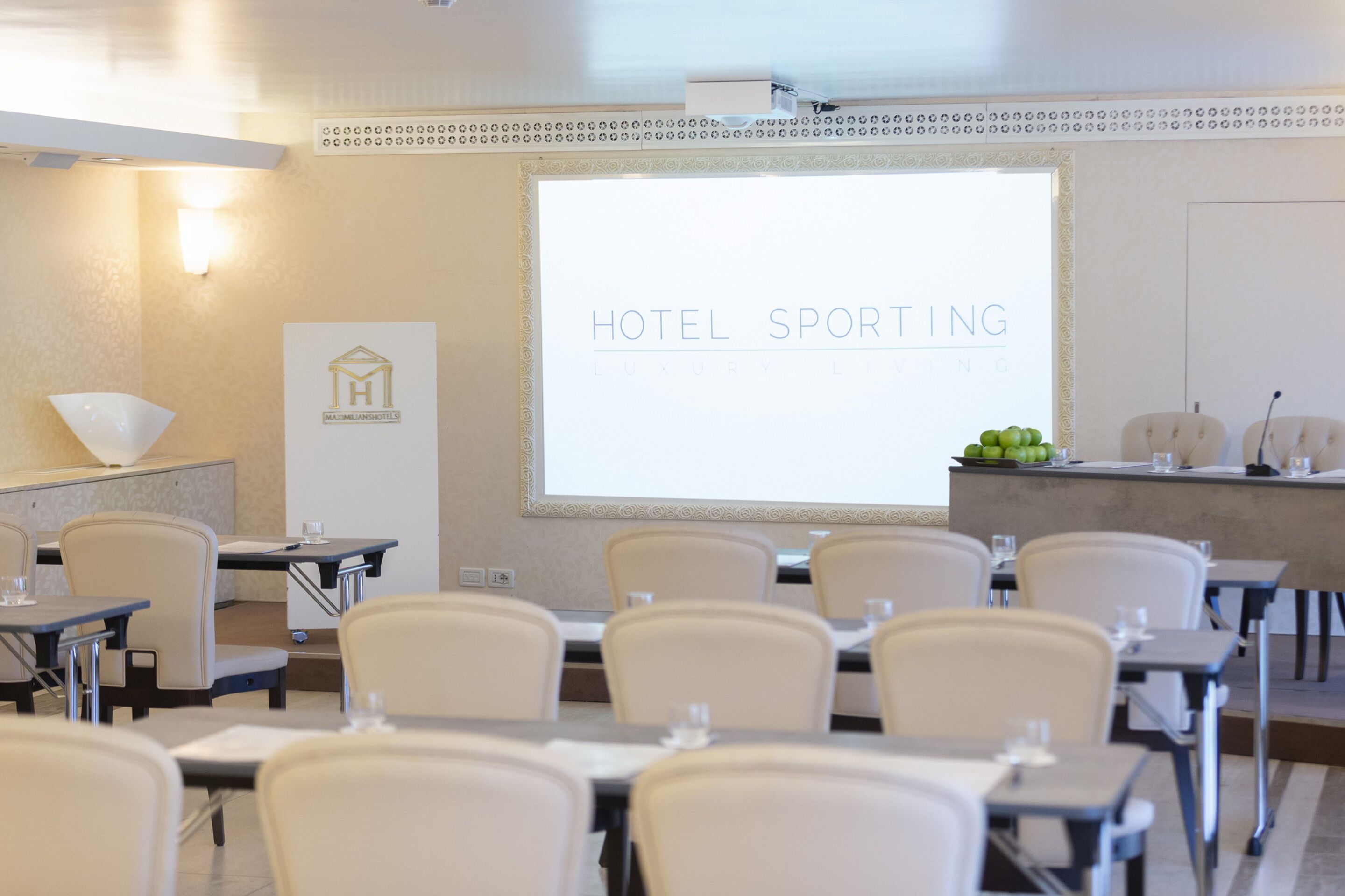 Et mødelokale med et projektionslærred, der viser "HOTEL SPORTING LUXURY LIVING".