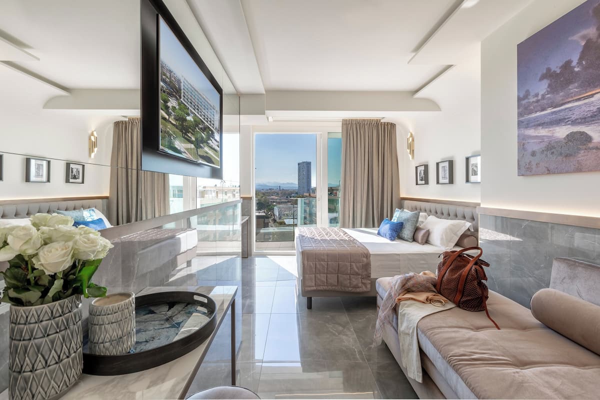 Ein modernes Hotelzimmer mit Balkon, einem großen Fernseher, einem Bett, einer Couch und einem Esstisch.